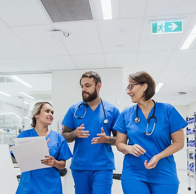 Two female nurses and one male nurse walking and talking in a hospital. One female nurse holds a file folder.